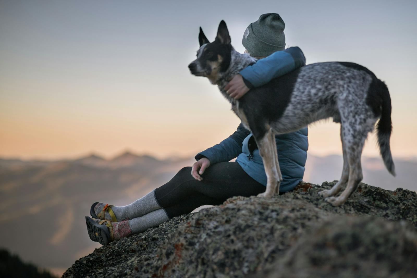 Photo by Bekka Mongeau A person wearing a beanie and winter jacket sits on a rocky mountain with a dog, overlooking sunset views.