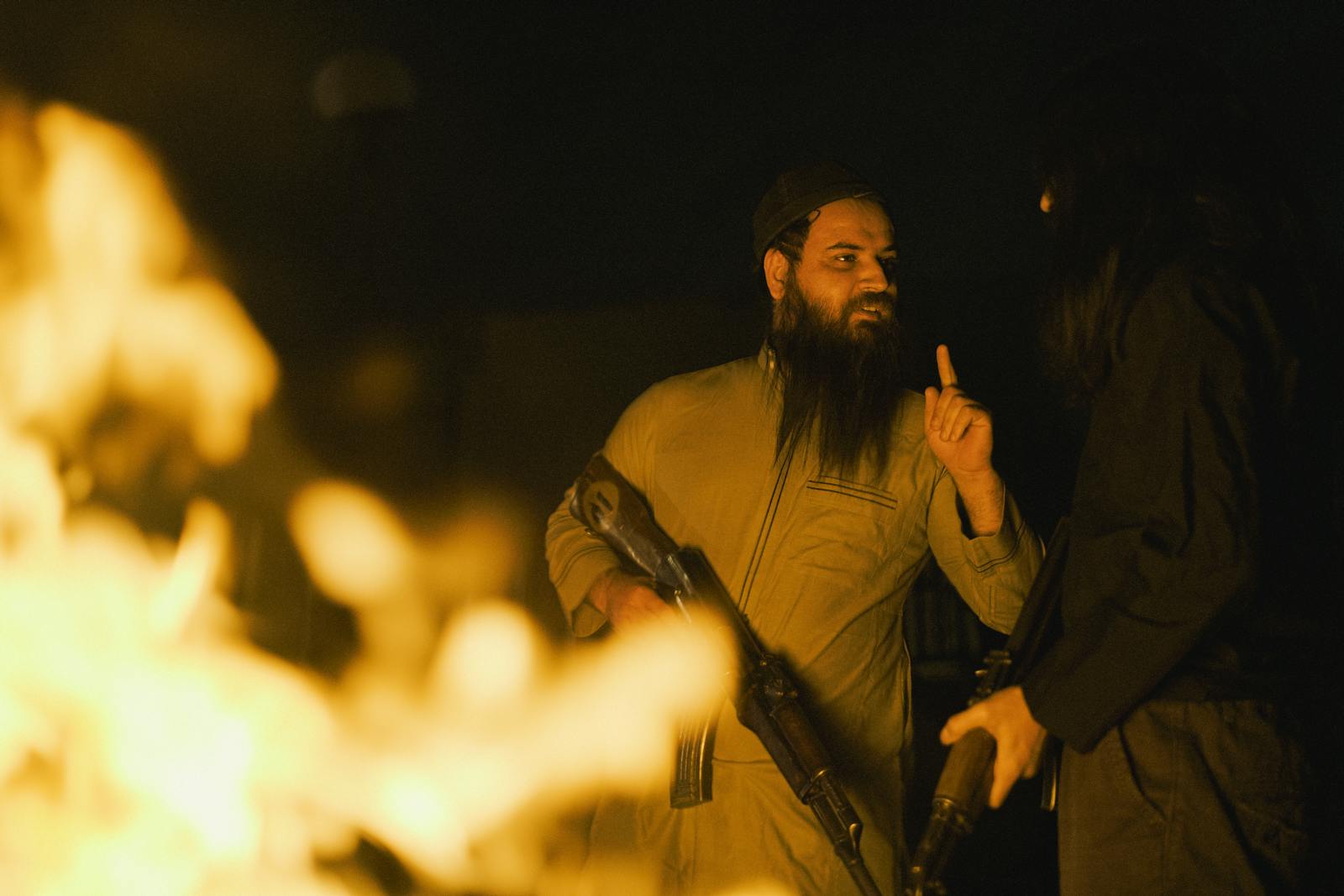Photo by AMORIE SAM Two men engage in a serious conversation beside a campfire at night, holding rifles.