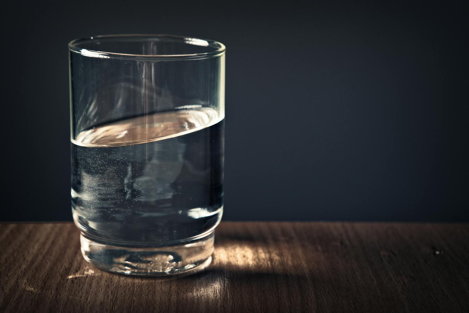 Photo by Stephan Müller A clear glass of water sitting on a wooden table against a dark background.