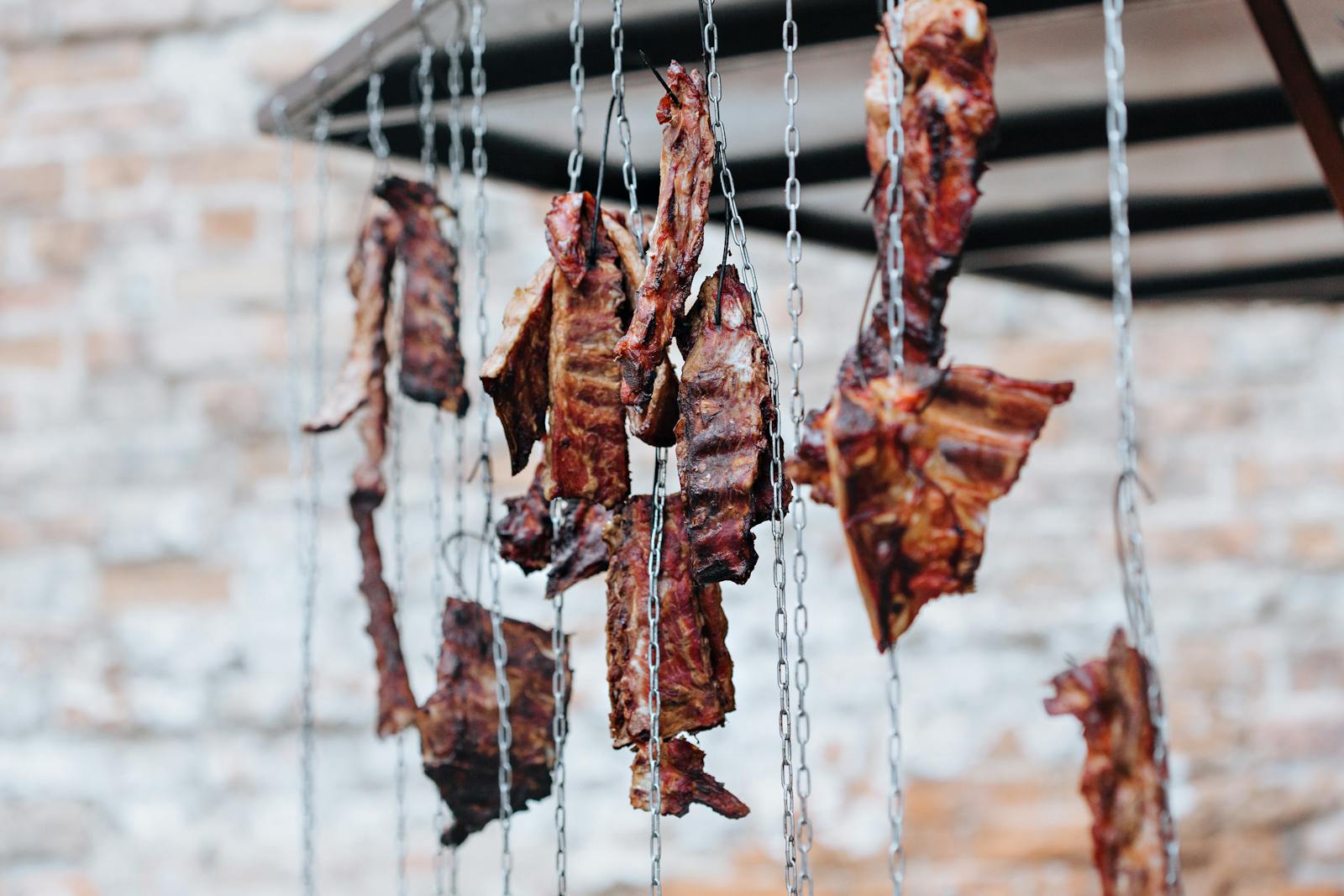 Photo by Julia Filirovska Rusted chains holding dried meat slices hanging against a brick backdrop.