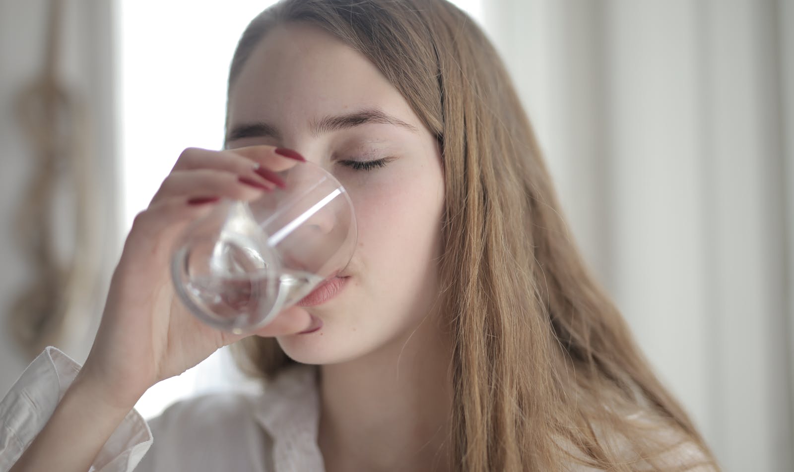 Photo by Andrea Piacquadio A young woman with brown hair drinks water from a clear glass indoors, eyes closed.