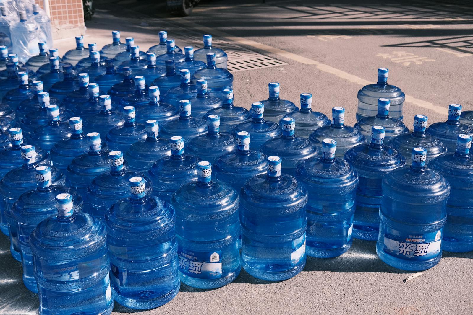 Photo by Junchen Zhou A collection of blue plastic water jugs lined up on an urban street in sunlight.