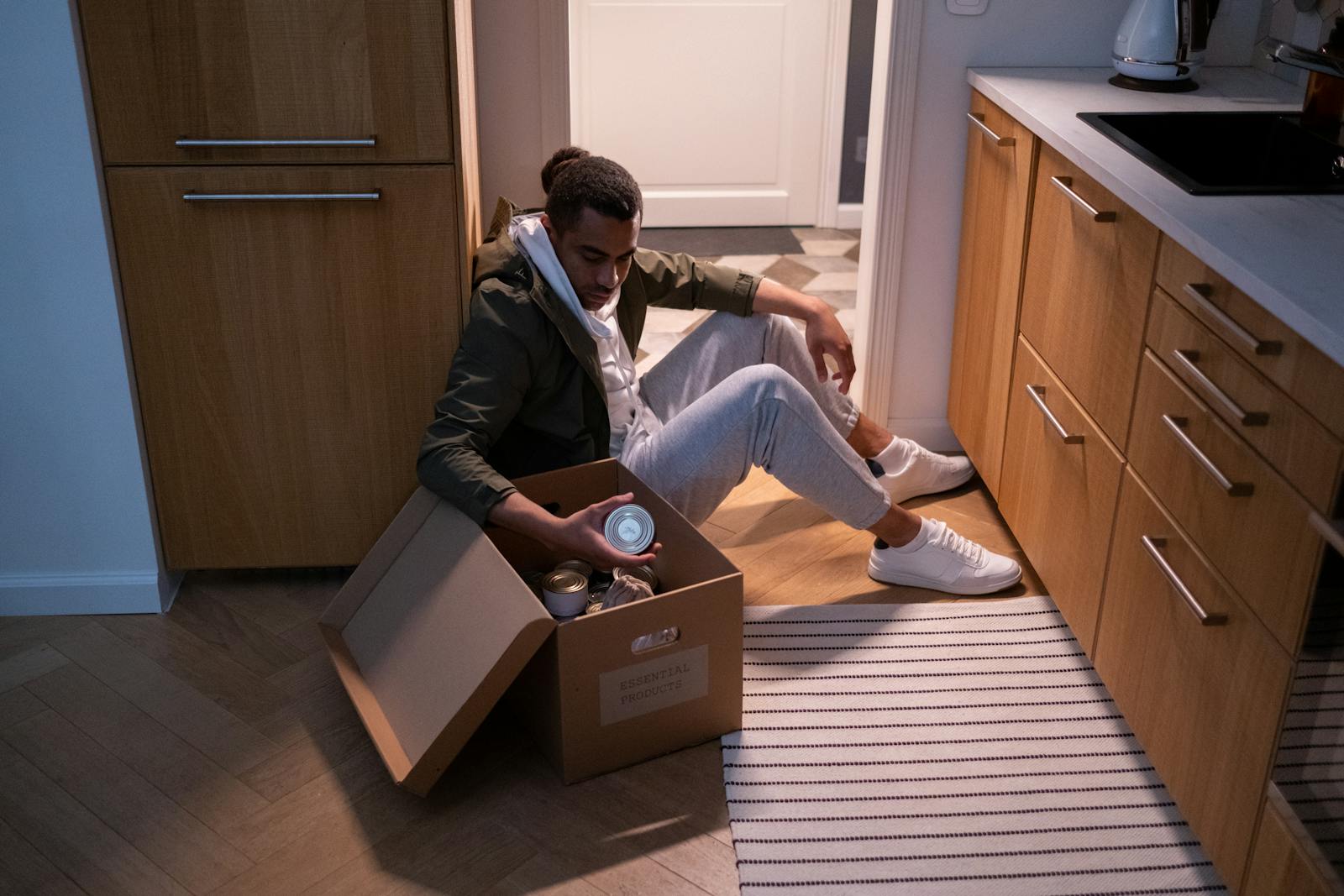 A man sits on the kitchen floor with an open box of canned goods, captured in a cozy indoor setting.