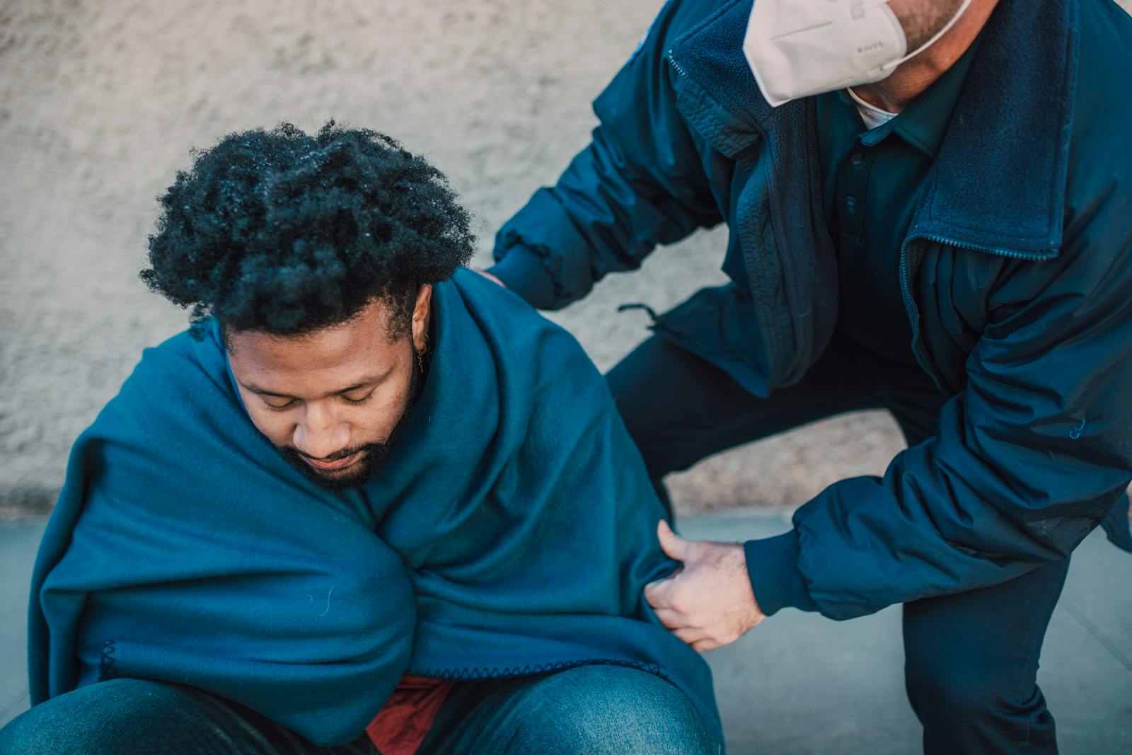 A paramedic assists an adult man covered with a blanket outdoors, showing care and support.